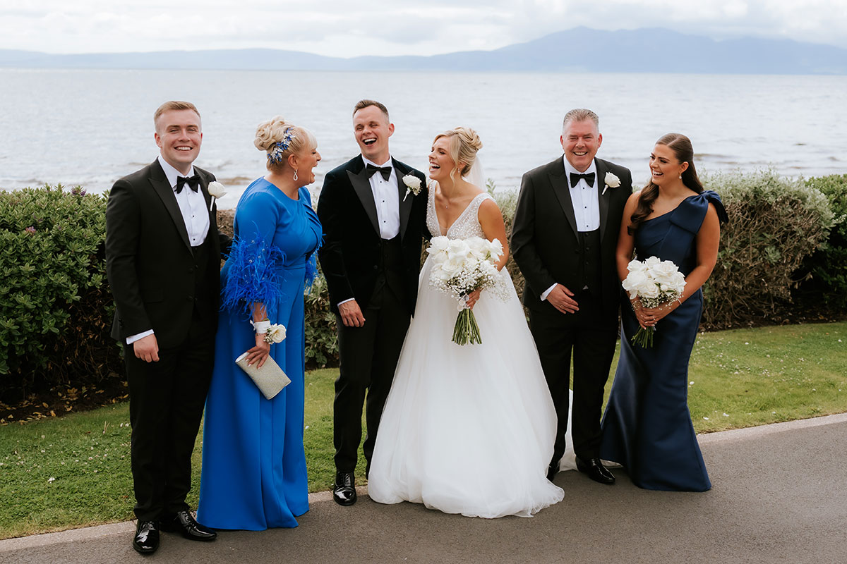 Bride Nicole and groom stand outdoors with family members by the coast, all dressed in formal wedding attire, with the sea and distant hills forming a scenic backdrop