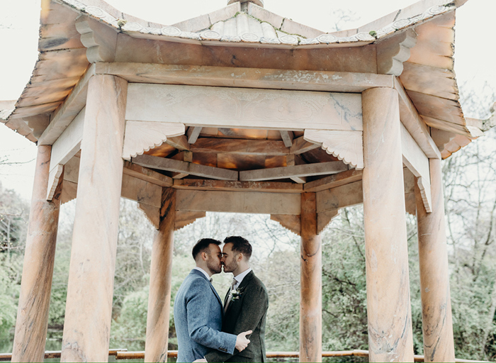 two grooms kissing under a wooden pagoda structure surrounded by green woodland