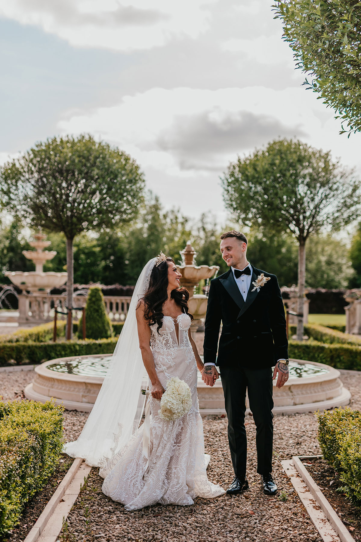 bride and groom smile at one another as they walk hand in hand through garden at ingliston