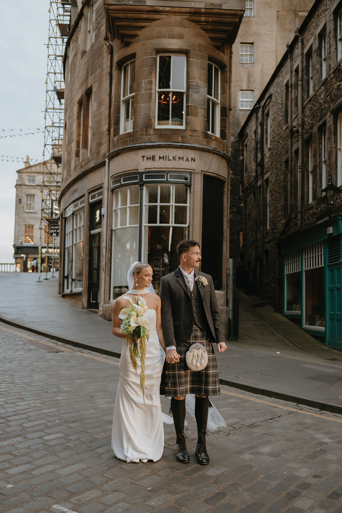 Bride and groom in Scottish wedding attire posing on a cobbled street beside The Milkman in Edinburgh Old Town