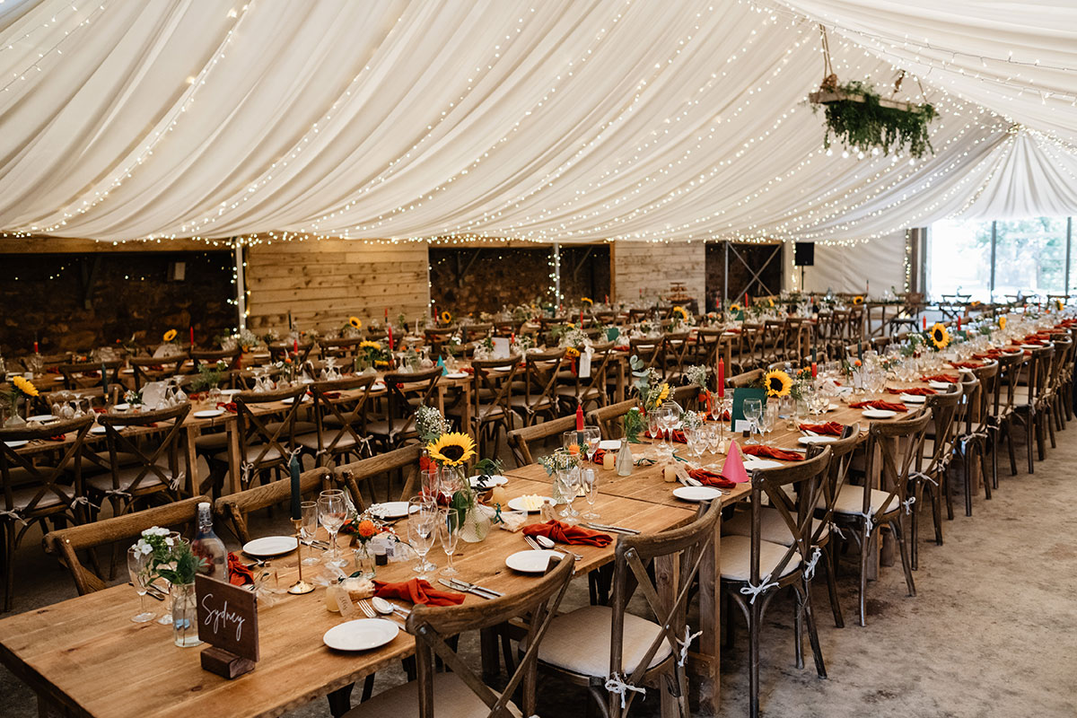 Rustic wedding reception setup with long wooden tables, sunflower centrepieces and draped marquee ceiling lights