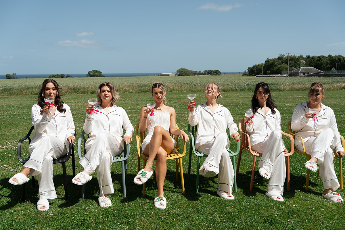Bride and bridesmaids relax outdoors in matching white pyjamas while enjoying cocktails overlooking the countryside at Kinkell Byre in St Andrews.