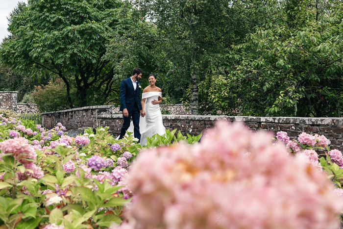 a joyous bride and groom walk next to a stone wall while pink and lilac hydrangeas blossom in the foreground