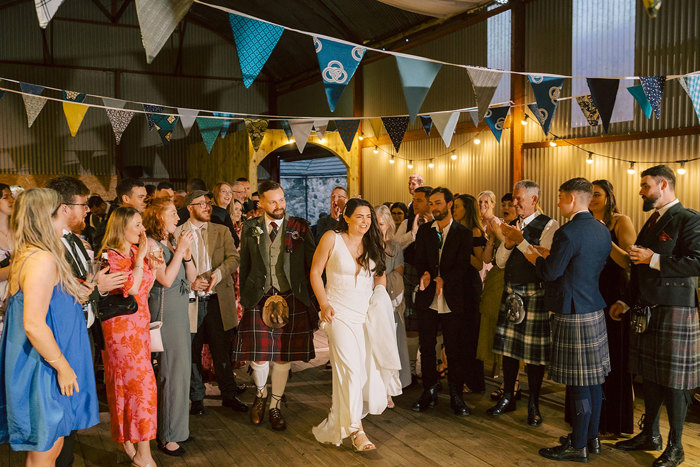 A bride and groom walking into a barn lit up with fairylights and decorated with bunting as their guests gather around and clap