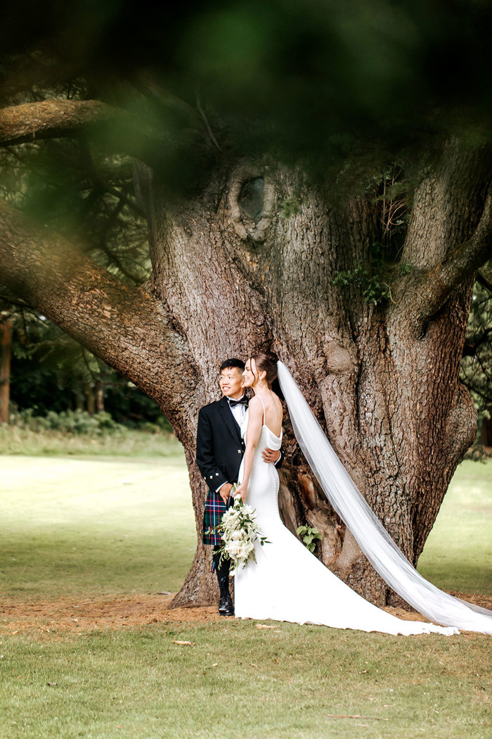 bride and groom wearing wedding outfits stand against tree at mar hall