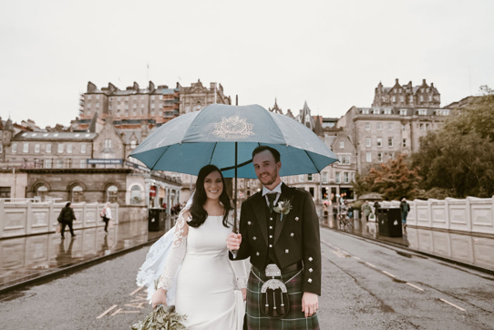 Groom holds umbrella over bride as they walk through Edinburgh