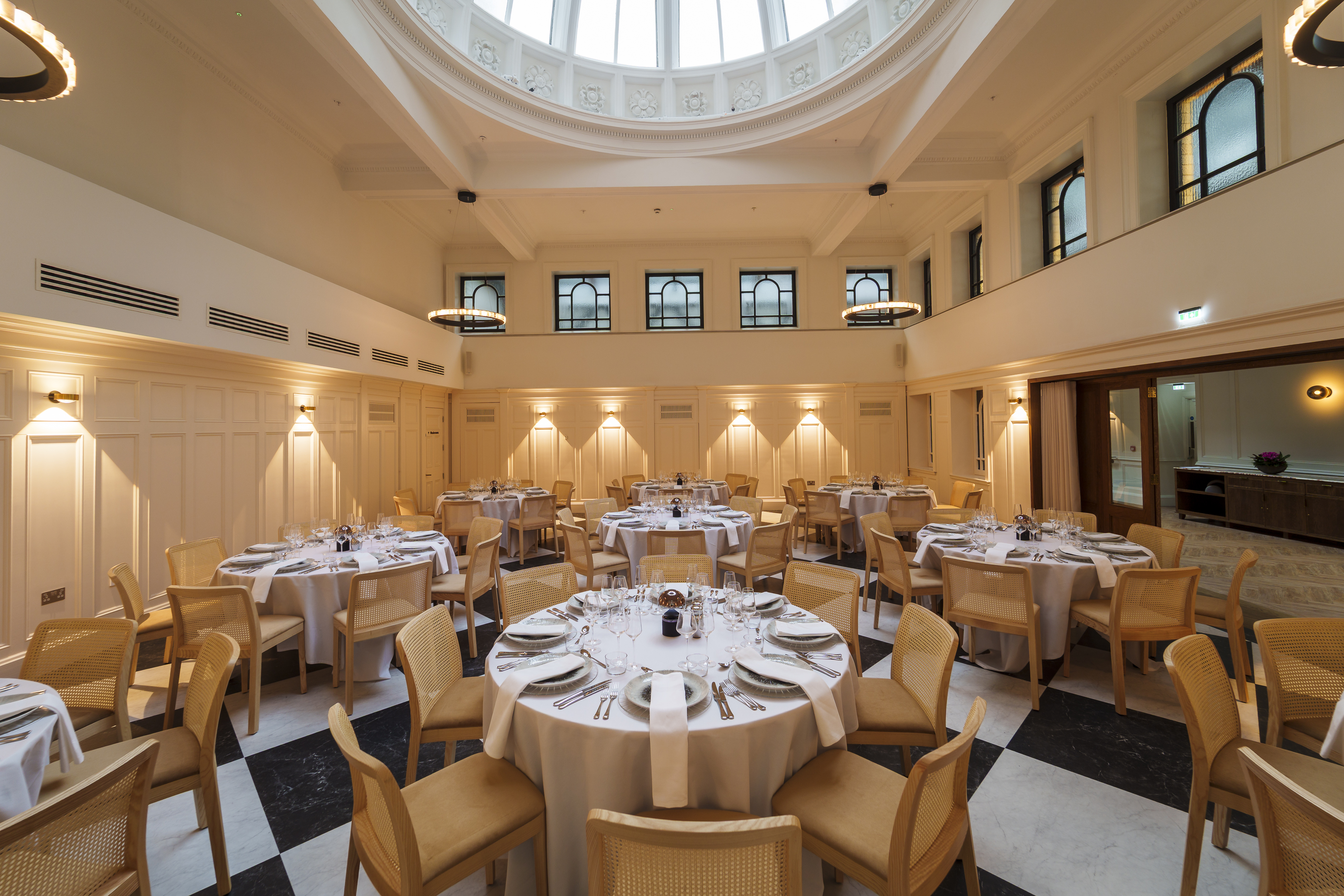 round tables with white linen and tableware set up for a wedding dinner below a glass dome on the ceiling