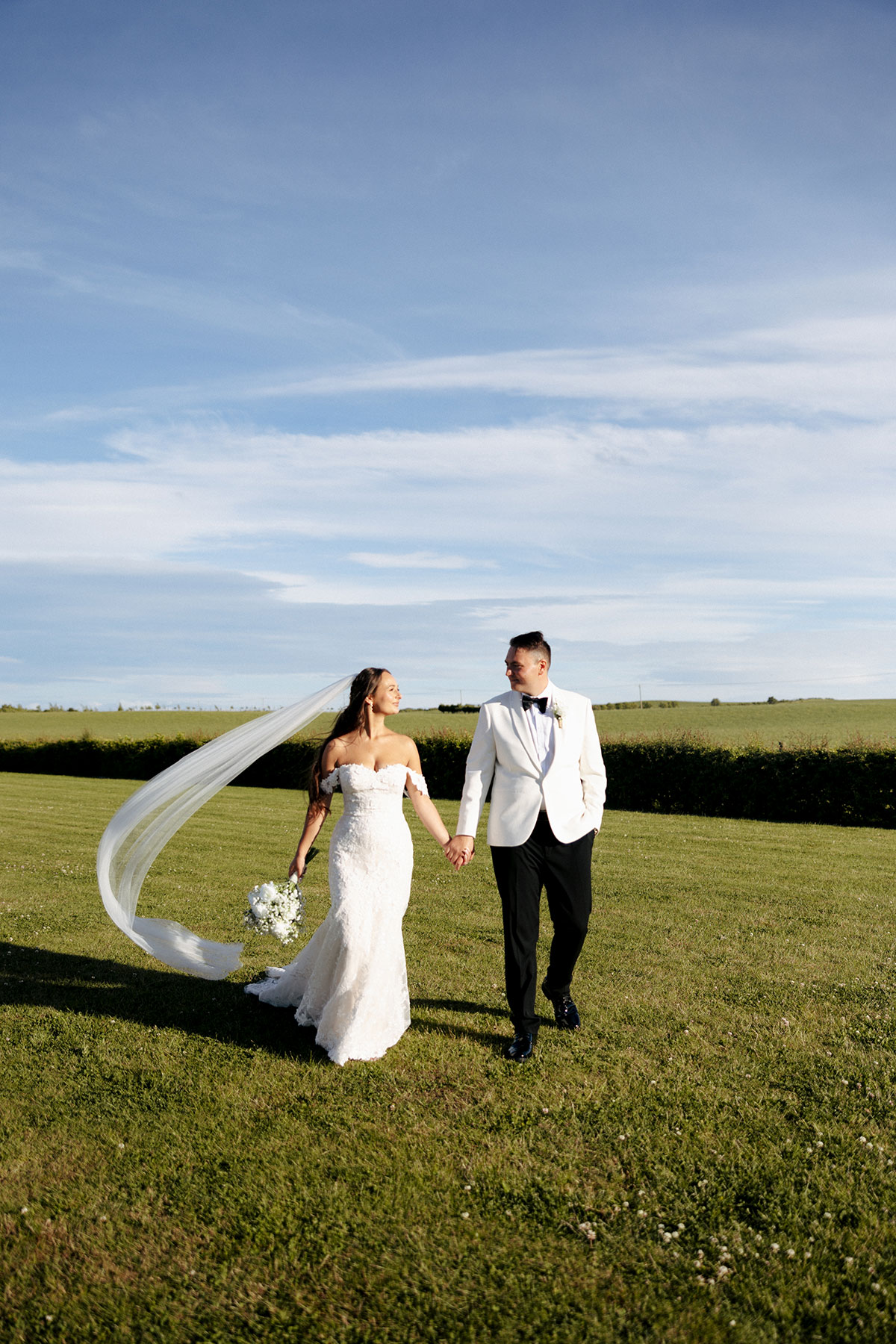 Bride and groom walk hand in hand across a green field at Falside Mill, the bride’s long veil flowing in the breeze.