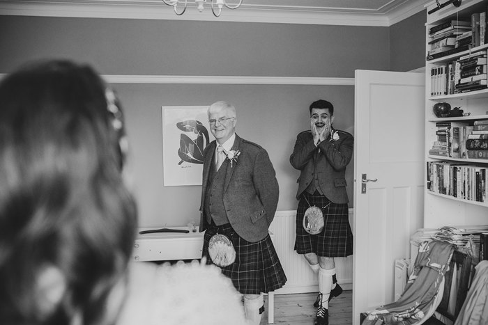 Black And White Image Of Two Men In Kilts Looking Surprised As They Look At A Bride