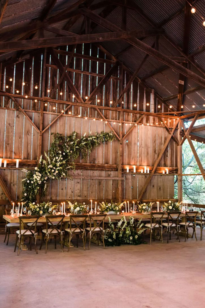 inside a barn with a long wooden table with greenery and white flowers on it and candles and a piece of greenery draped on the wall behind