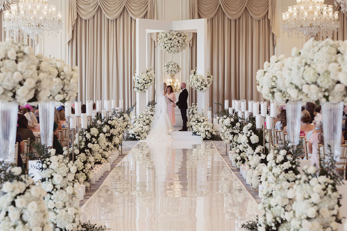 Luxury indoor wedding ceremony setup with white floral aisle and mirrored walkway