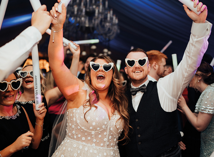 A bride and groom standing close to the camera with a crowd of people behind them, all wearing white heart shaped sunglasses and holding light up batons