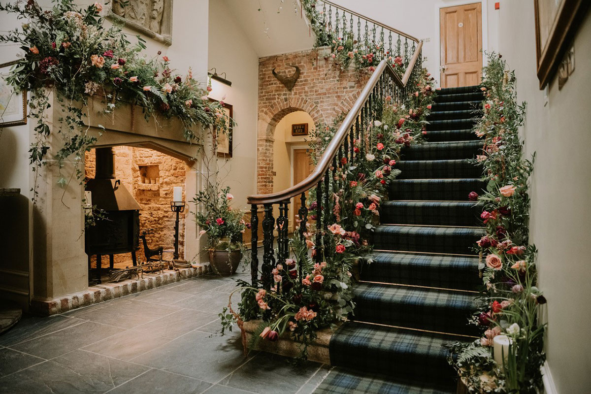A tartan-carpeted staircase lined with lush floral arrangements, next to a stone fireplace.