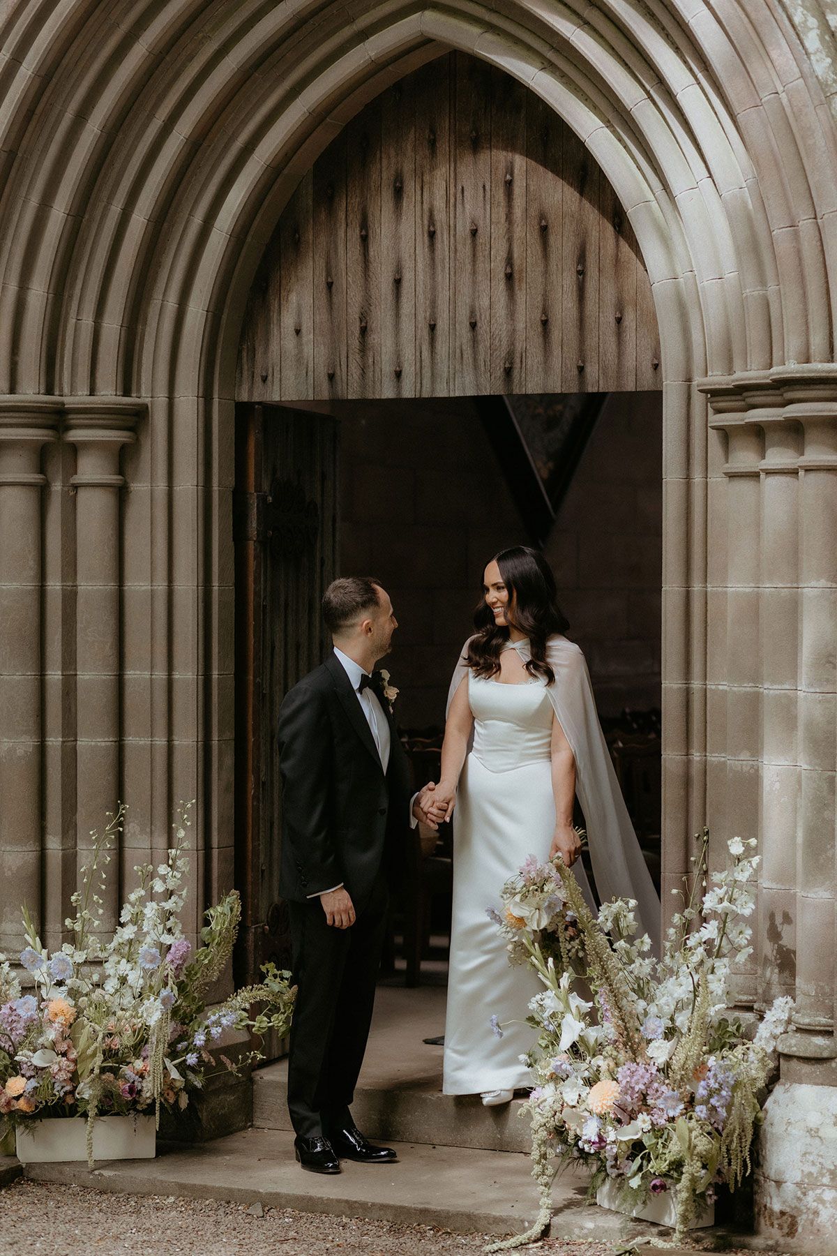 Newlyweds holding hands in chapel doorway at Rosebery House and Steading, Midlothian, framed by floral ceremony arrangements.