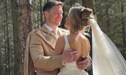 a bride wearing a wedding dress and a groom wearing a beige kilt stand with their arms around each other in the woodlands