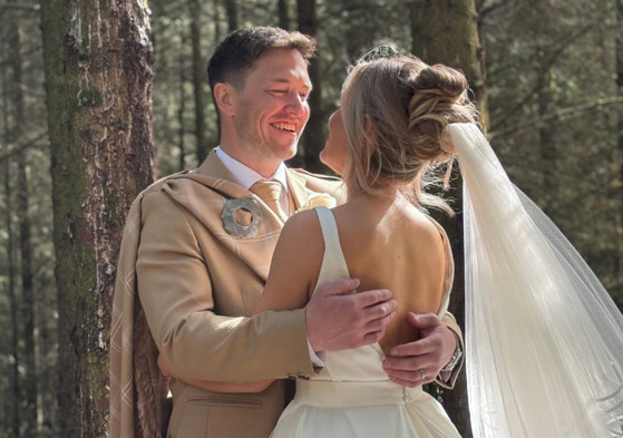 a bride wearing a wedding dress and a groom wearing a beige kilt stand with their arms around each other in the woodlands