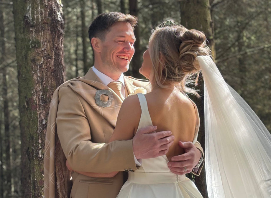 a bride wearing a wedding dress and a groom wearing a beige kilt stand with their arms around each other in the woodlands