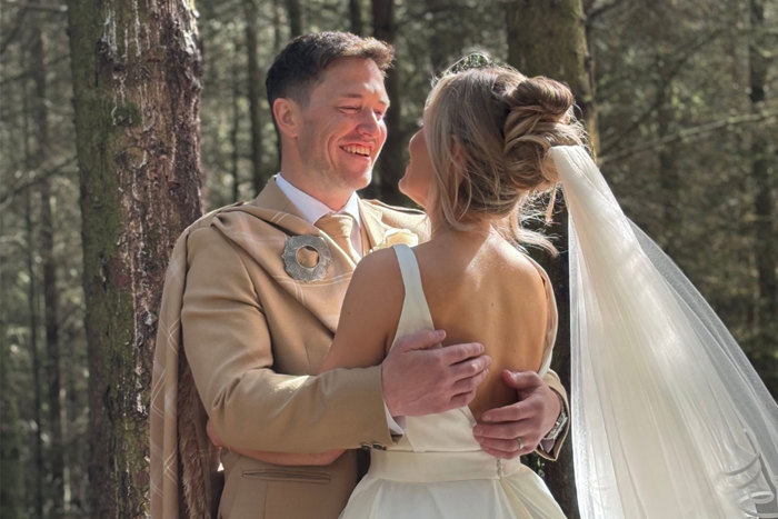a bride wearing a wedding dress and a groom wearing a beige kilt stand with their arms around each other in the woodlands