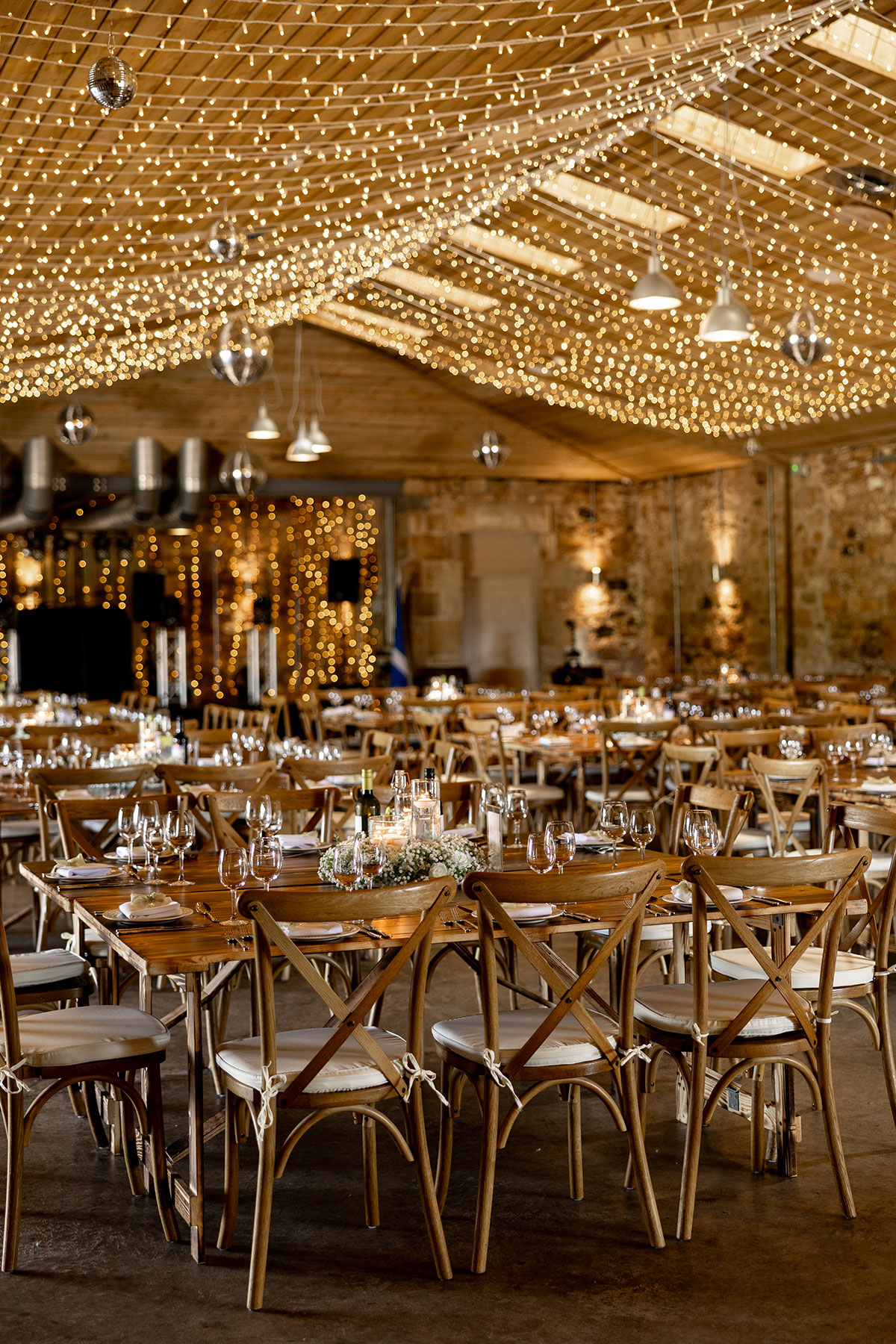Falside Mill tables set for a wedding dinner with wooden cross back chairs. The softly lit barn venue is decorated with candles and white flowers.