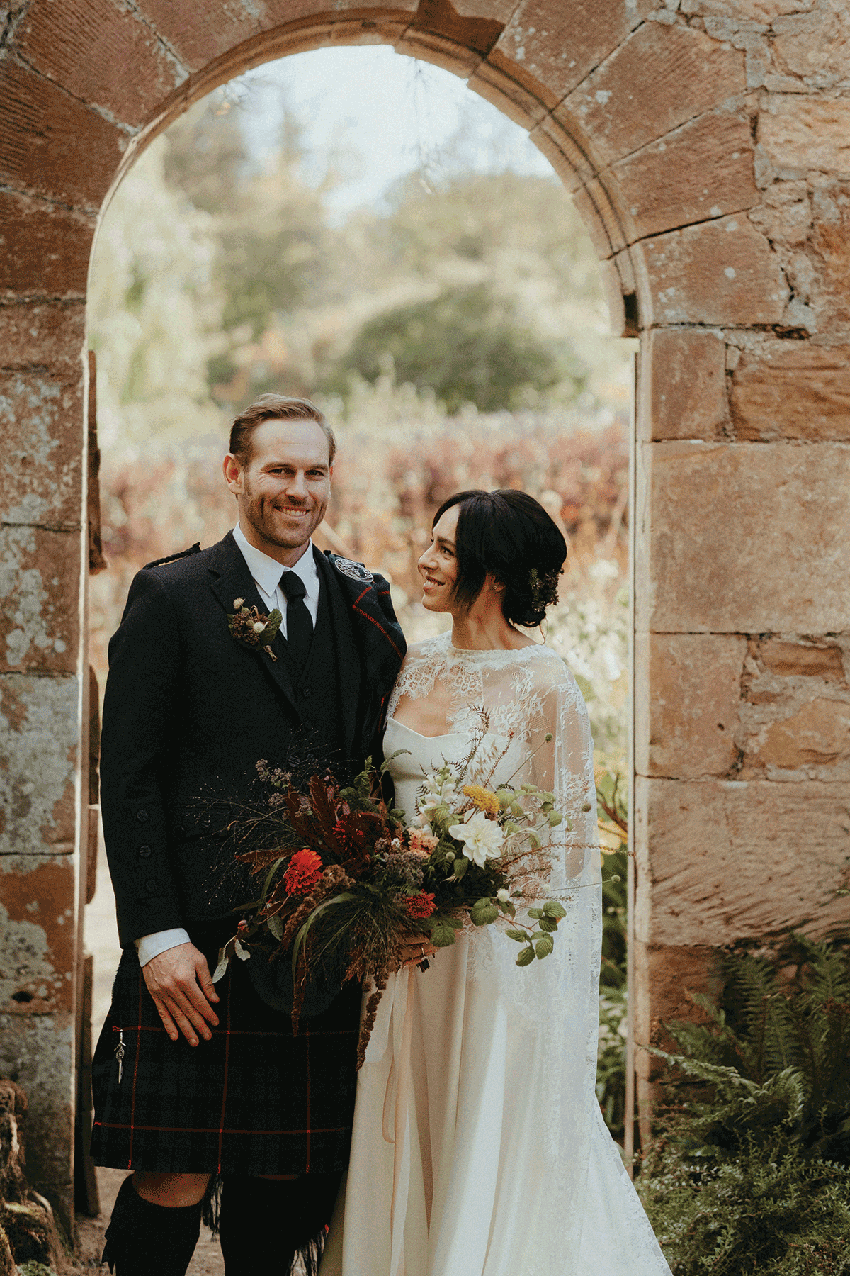 Bride and groom standing beneath a stone archway in a historic Scottish garden, featuring romantic florals, Highlandwear and autumn-inspired wedding styling