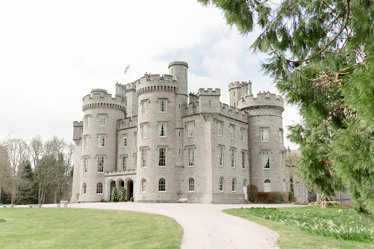 Grand stone castle with turrets and arched entrance, set within landscaped grounds and trees