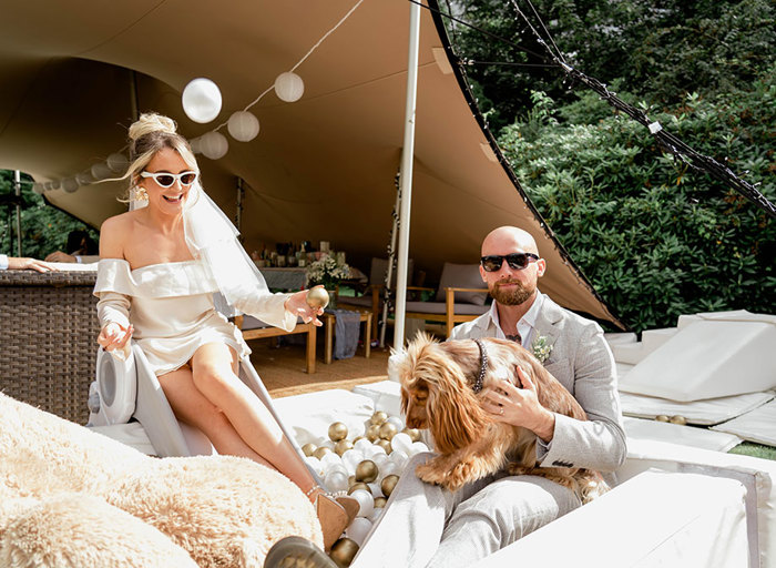 a bride, groom and dog sit on a white ball pit with canvas stretch tent in background