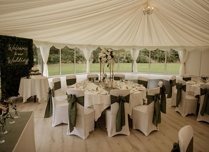 Round tables with white tablecloths and chair covers with green bows tied around the chairs inside a white marquee