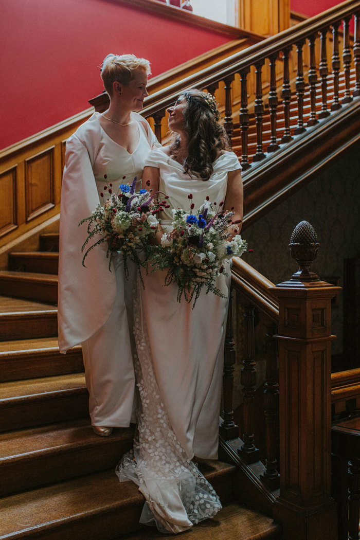 two brides holding wildflower bouquets as they stand on staircase in cambo house