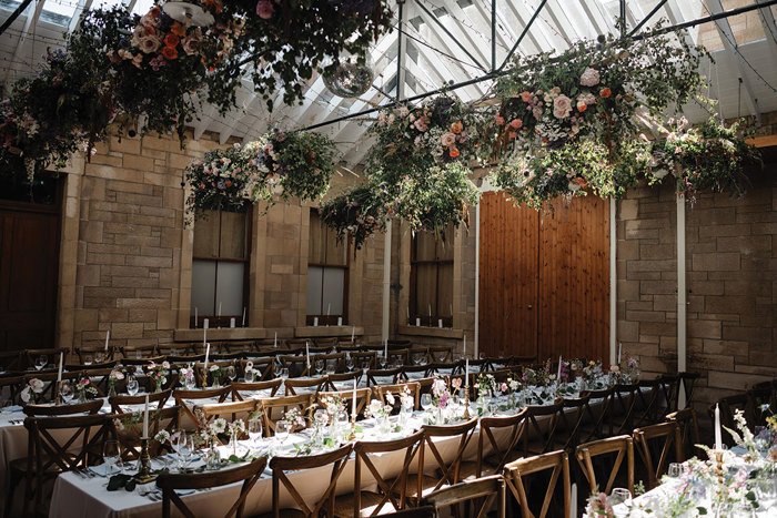 three rows of long wooden tables with place settings laid out and large floral cloud arrangements hanging overhead