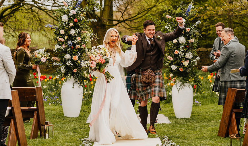 An elated newlywed bride and groom cheer and hold hands aloft in a lush outdoor ceremony setting in a garden. The bride is holding a large pastel multicolour bouquet with trailing ribbon and flowing ivory long sleeved dress. Two large white urns are behind the couple, filled with tall vertical flower arrangements. Guests look on, standing up from wooden chairs, and cheer and clao