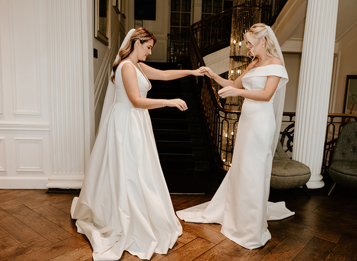 Two brides holding hands and admiring one another's wedding dresses at the bottom of a staircase at Kimpton Blythswood Square Hotel