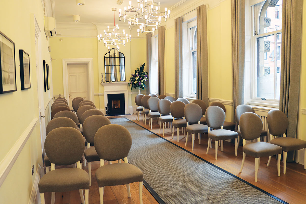 Ceremony room set for a small wedding at Glasgow Registrars Office with chairs arranged beside large windows and a fireplace.
