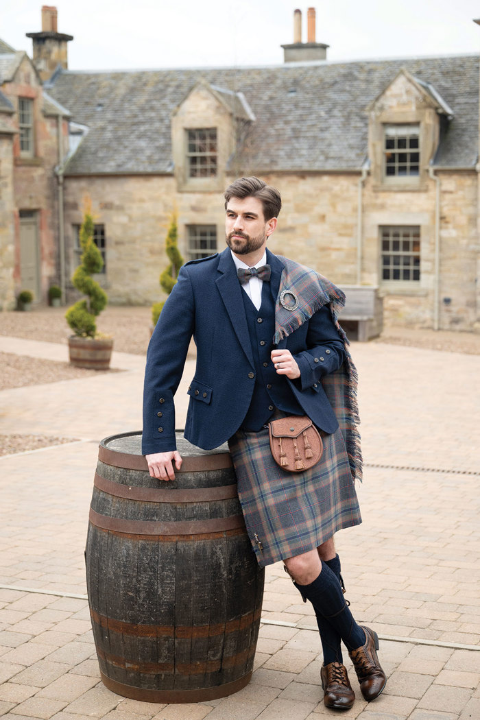 A man standing leaning against a whisky barrel in the courtyard at Rosebery Steading. He is wearing a kilt outfit from MacGregor and MacDuff.