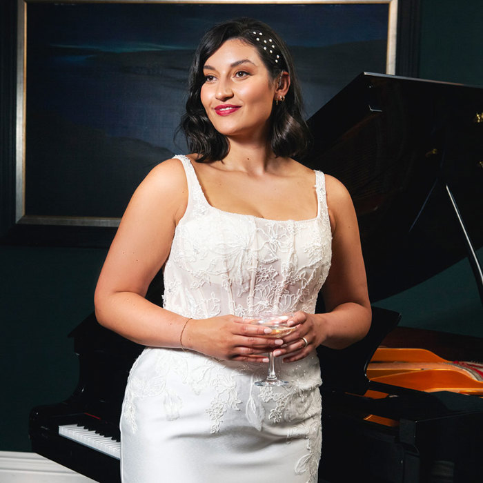 A woman wearing a white fitted dress holds a coupe of champagne and leaning against a piano