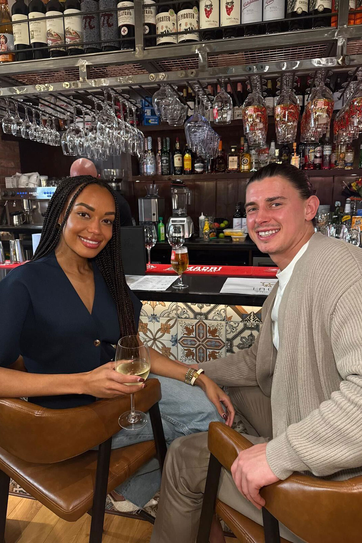 A smiling couple sit together at a bar, each holding a glass of wine, with shelves of bottles and hanging glassware behind them