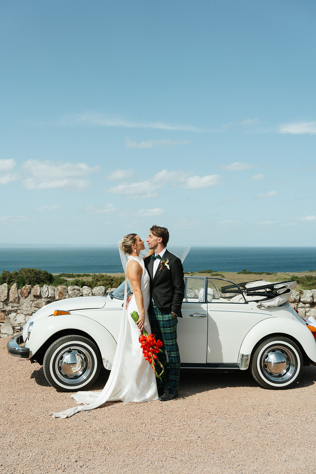 Bride and groom pose beside a white vintage Volkswagen Beetle convertible with coastal views behind them at Kinkell Byre in St Andrews.