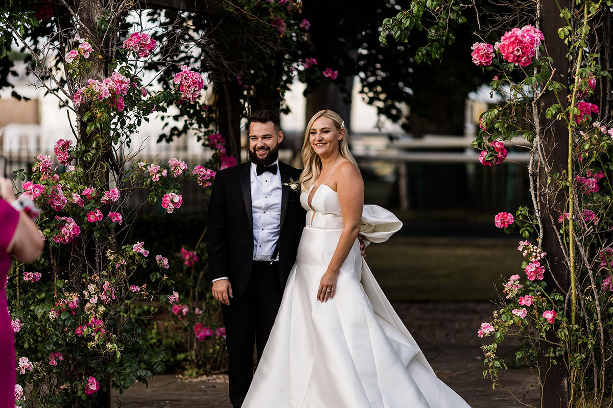Bride and groom standing beneath pink rose-covered arches in the gardens at Western House Hotel, Ayr.