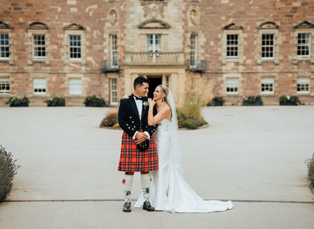 A bride in a wedding dress and a groom in a kilt standing in front of Archerfield House.