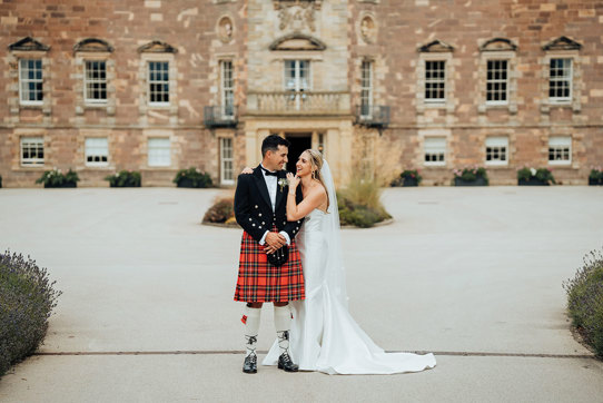 A bride in a wedding dress and a groom in a kilt standing in front of Archerfield House.