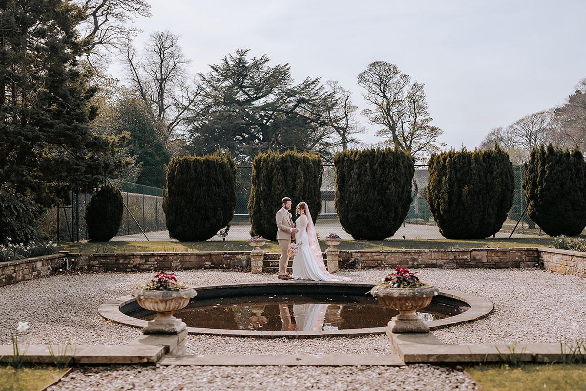 a bride and groom posing by an ornamental pond and hedges in the garden at Carlowrie Castle Edinburgh wedding venue