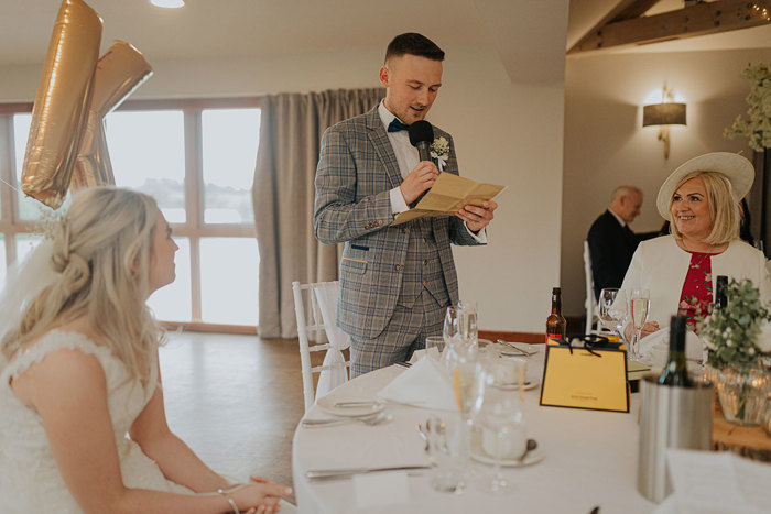 A Groom Making A Speech Standing At A Round Table As Bride And Guest Watch On