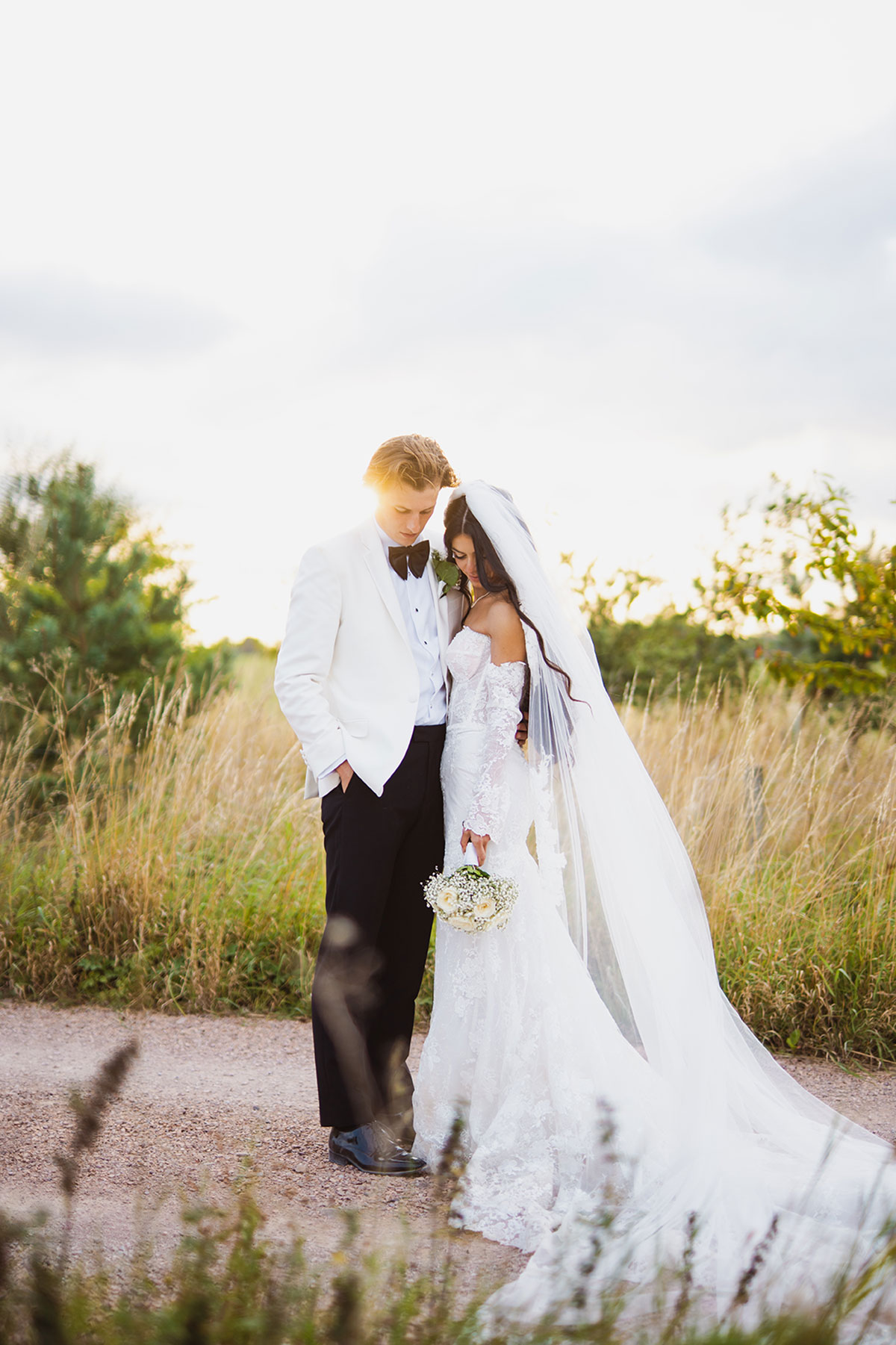 Bride and groom portrait at golden hour in long grass, wearing white tux jacket and lace wedding dress
