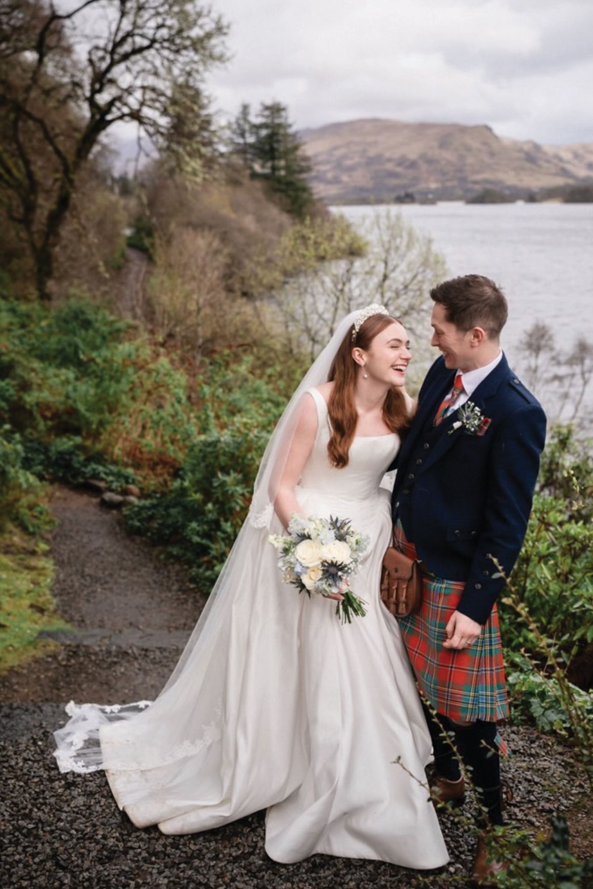 Scottish wedding couple by a loch, with the groom wearing a kilt and the bride holding a white and thistle bouquet.
