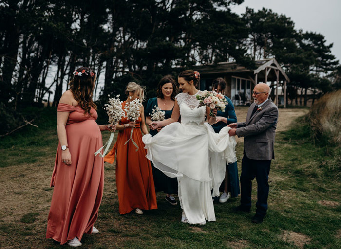 a bride being helped hold her skirt up by a group of people wearing wedding attire. There are tall trees and a wooden hut in the background and they stand on grassy and sandy ground