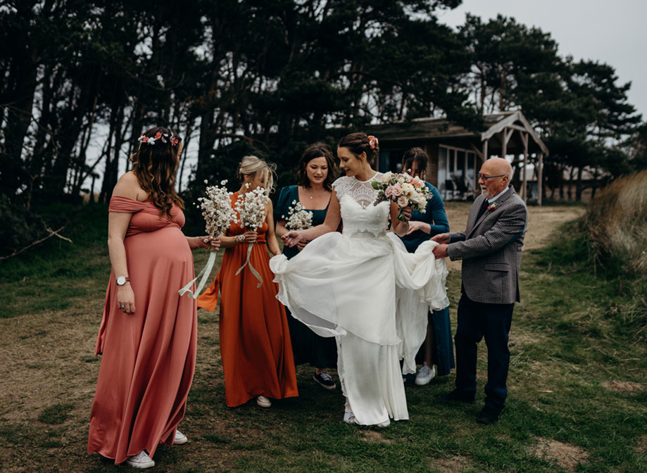 a bride being helped hold her skirt up by a group of people wearing wedding attire. There are tall trees and a wooden hut in the background and they stand on grassy and sandy ground