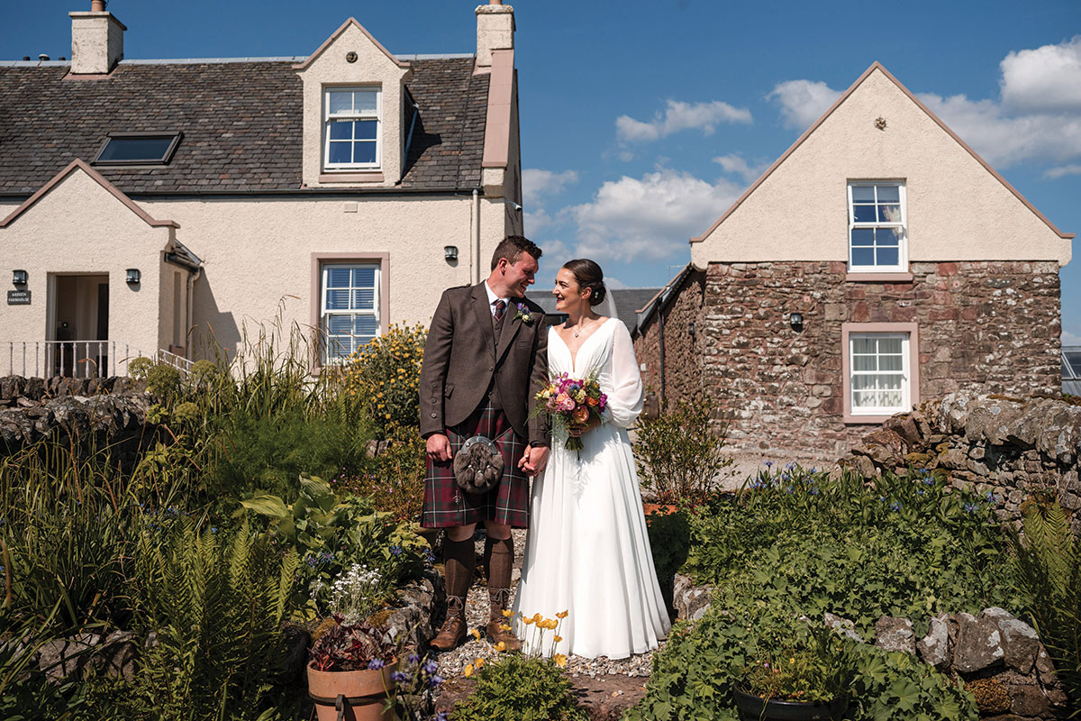 bride in long sleeve flowy wedding dress and groom in red kilt outfit stand side by side, looking into each other's eyes, in the midst of lots of planted and potted greenery, with two brick homes behind them