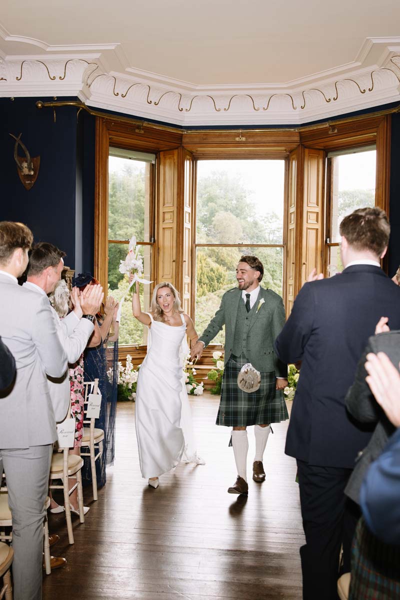 Lia and Louis walk back up the aisle as guests applaud following their wedding ceremony at Cambo House.
