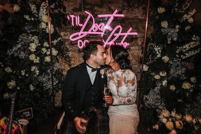 couple kissing on wedding day with sign that reads 'til death do us party' in the background