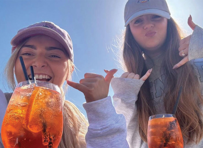 two women holding up thumbs in grey outfits and hats drinking wine glasses with orange liquid 