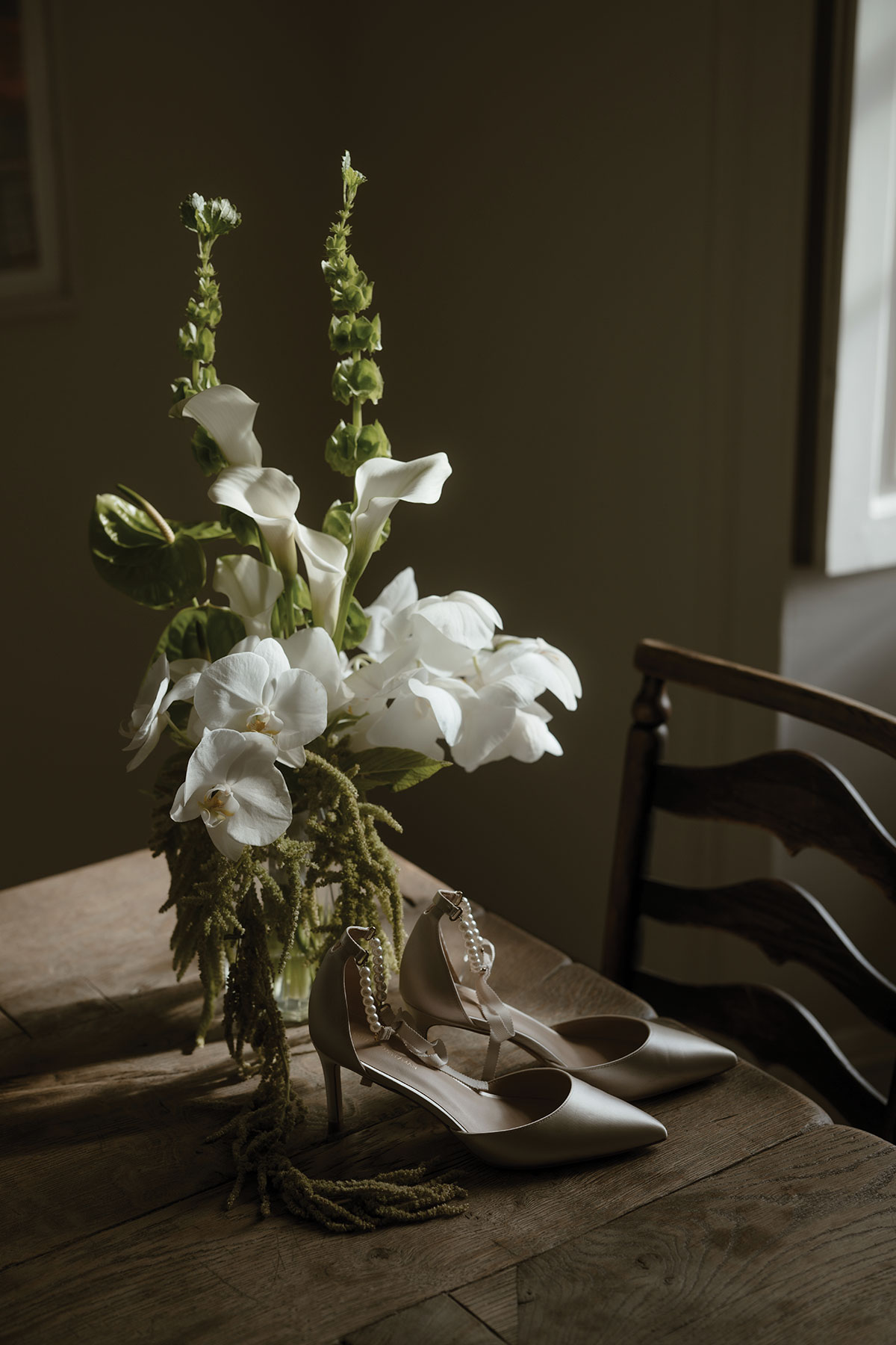Elegant white wedding bouquet with orchids and calla lilies arranged beside pearl-detail bridal heels on a wooden table